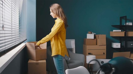 Junge Frau in gelber Bluse, die Kisten in einem Zimmer mit petrolfarbener Wand auspackt.